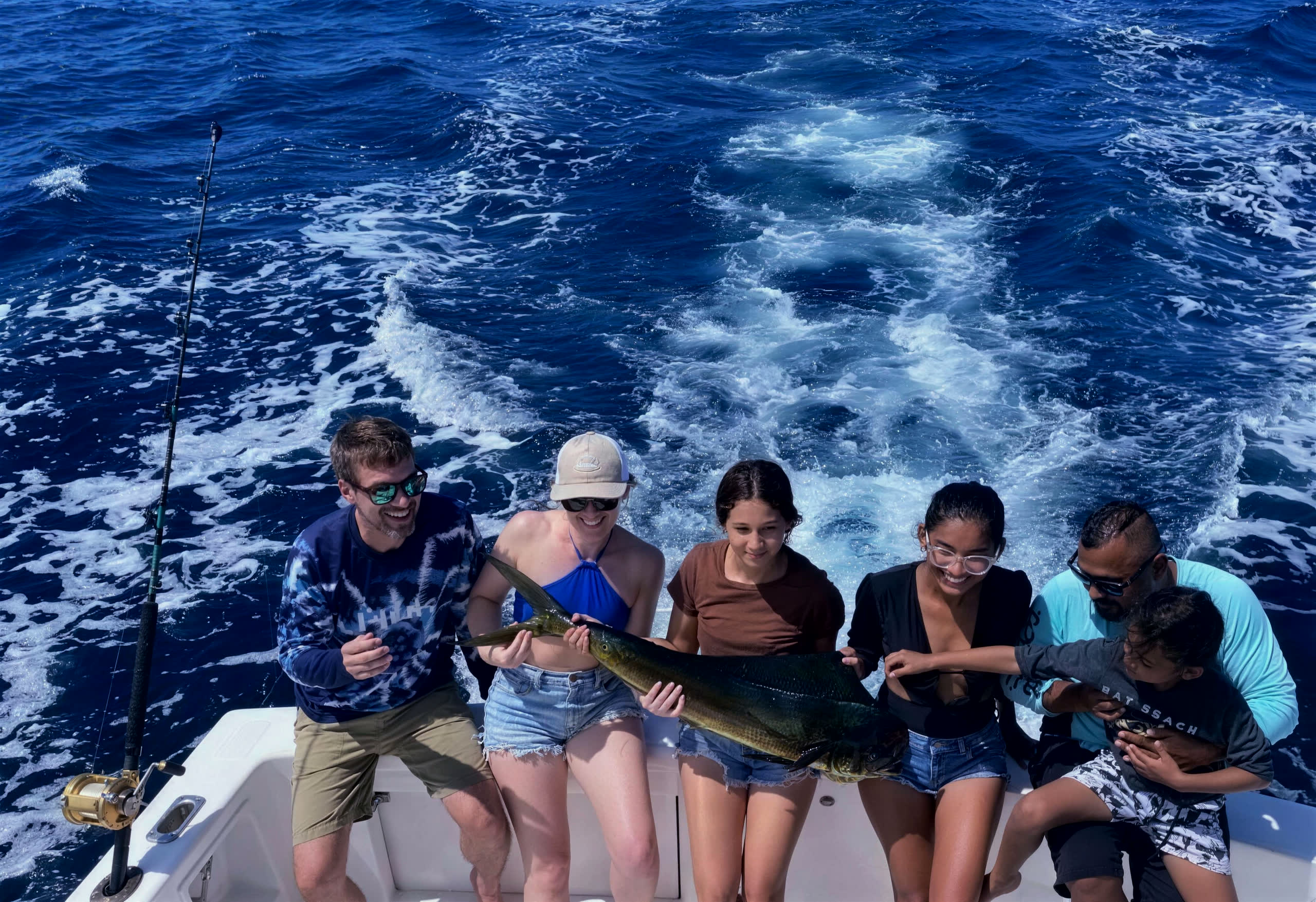 A family of six on the stern holding a mahi mahi, the wake bright behind them